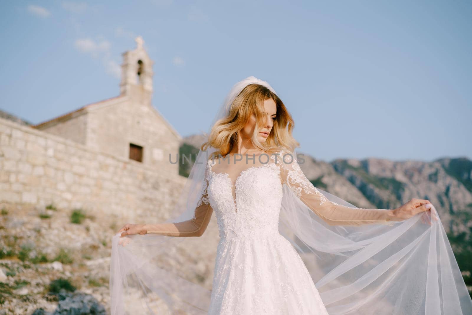 Bride in a white lace dress stands against the background of an ancient stone church by Nadtochiy