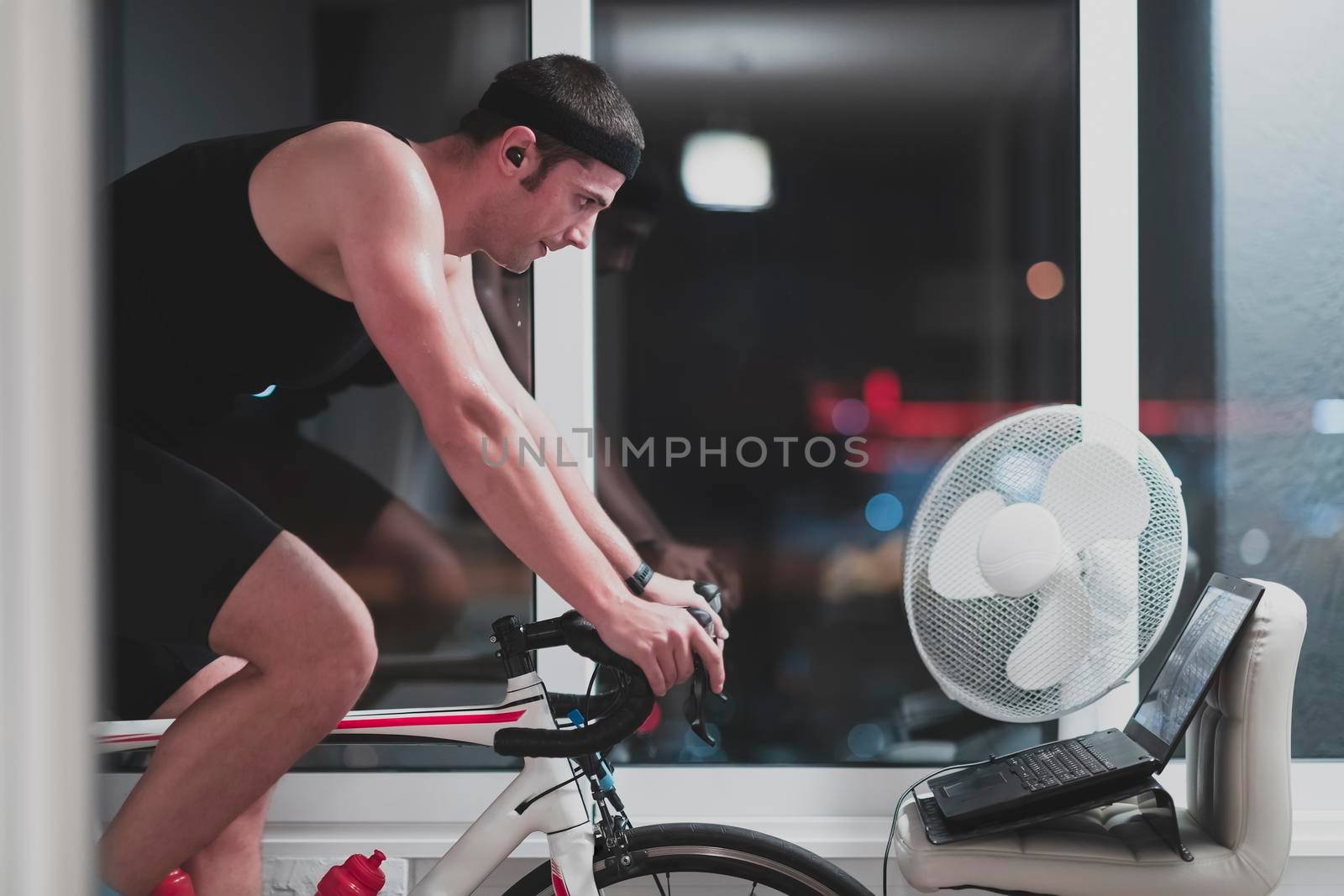 Man cycling on the machine trainer he is exercising in the home at night. Playing online bike racing game during coronavirus covid19 lockdown. New normal concept.