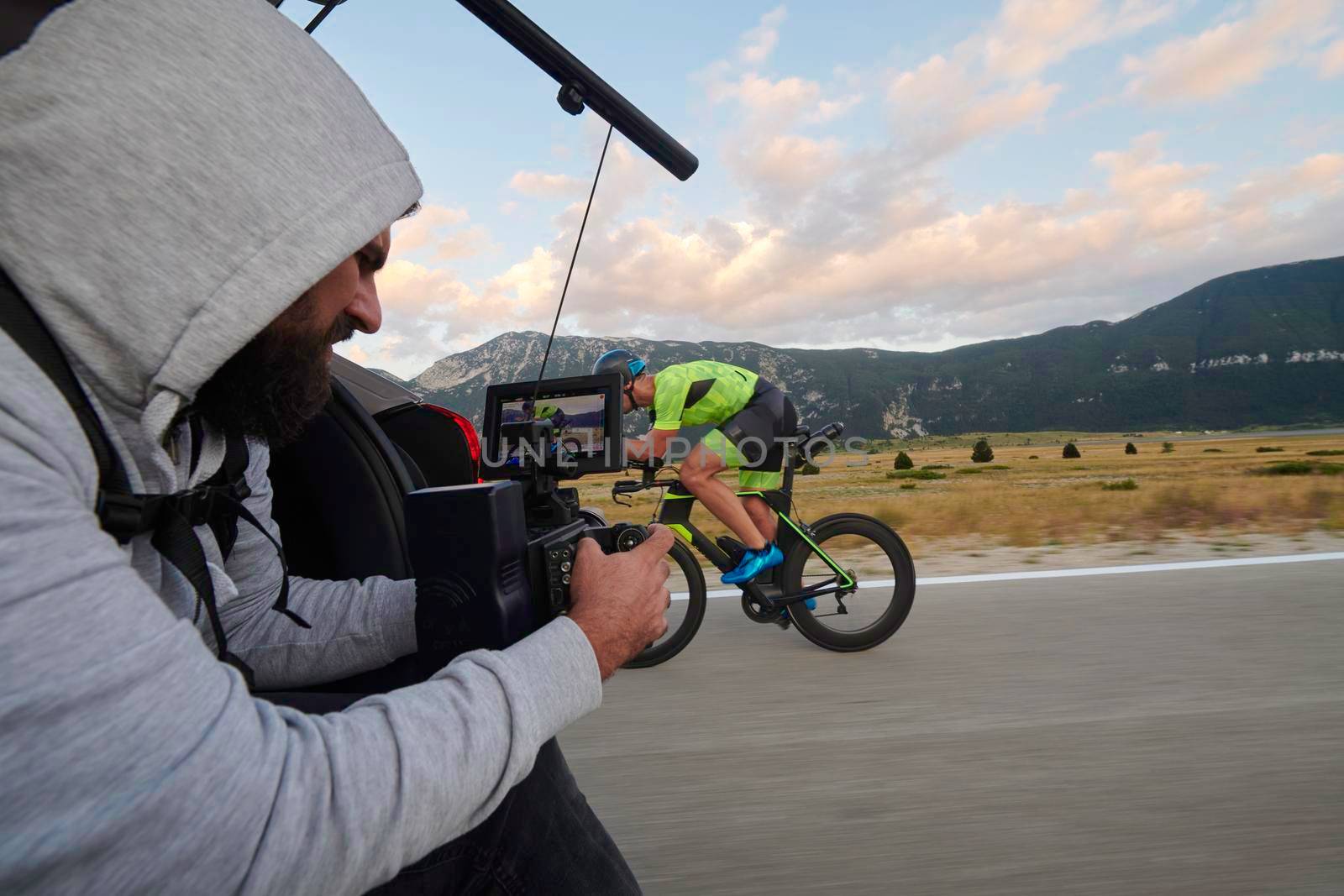 cinematographer with professional cinema camera taking sport action shot  from moving car of triathlon athlete who riding bike on low traffic country road