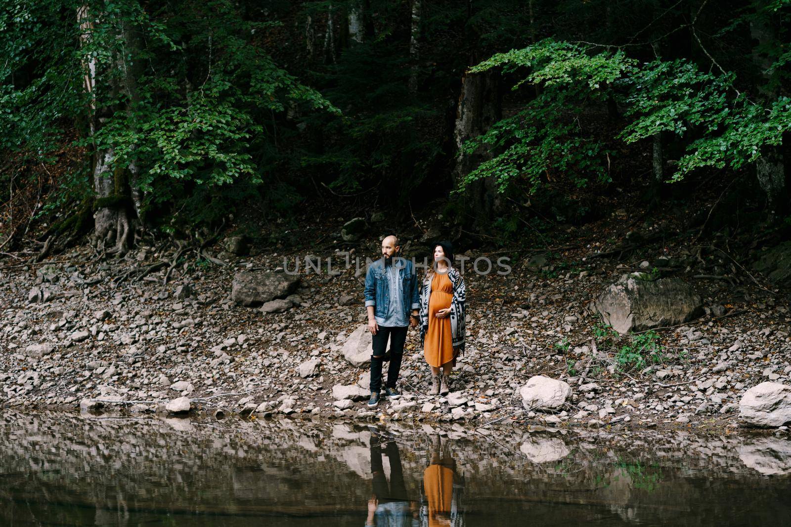 Pregnant woman with man stands on a rocky slope by a lake surrounded by trees. High quality photo