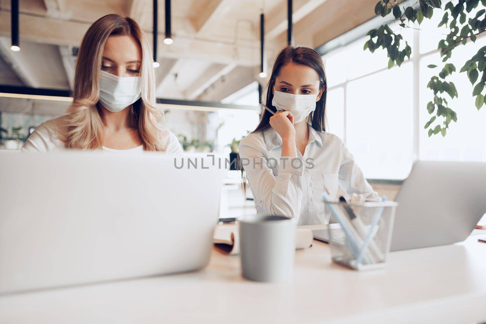 Two female colleagues working in office together wearing medical masks, coronavirus prevention