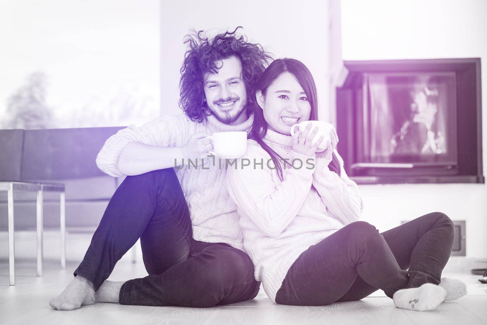 Young romantic multiethnic couple sitting on the floor in front of fireplace at home, talking and drinking coffee at cold winter day