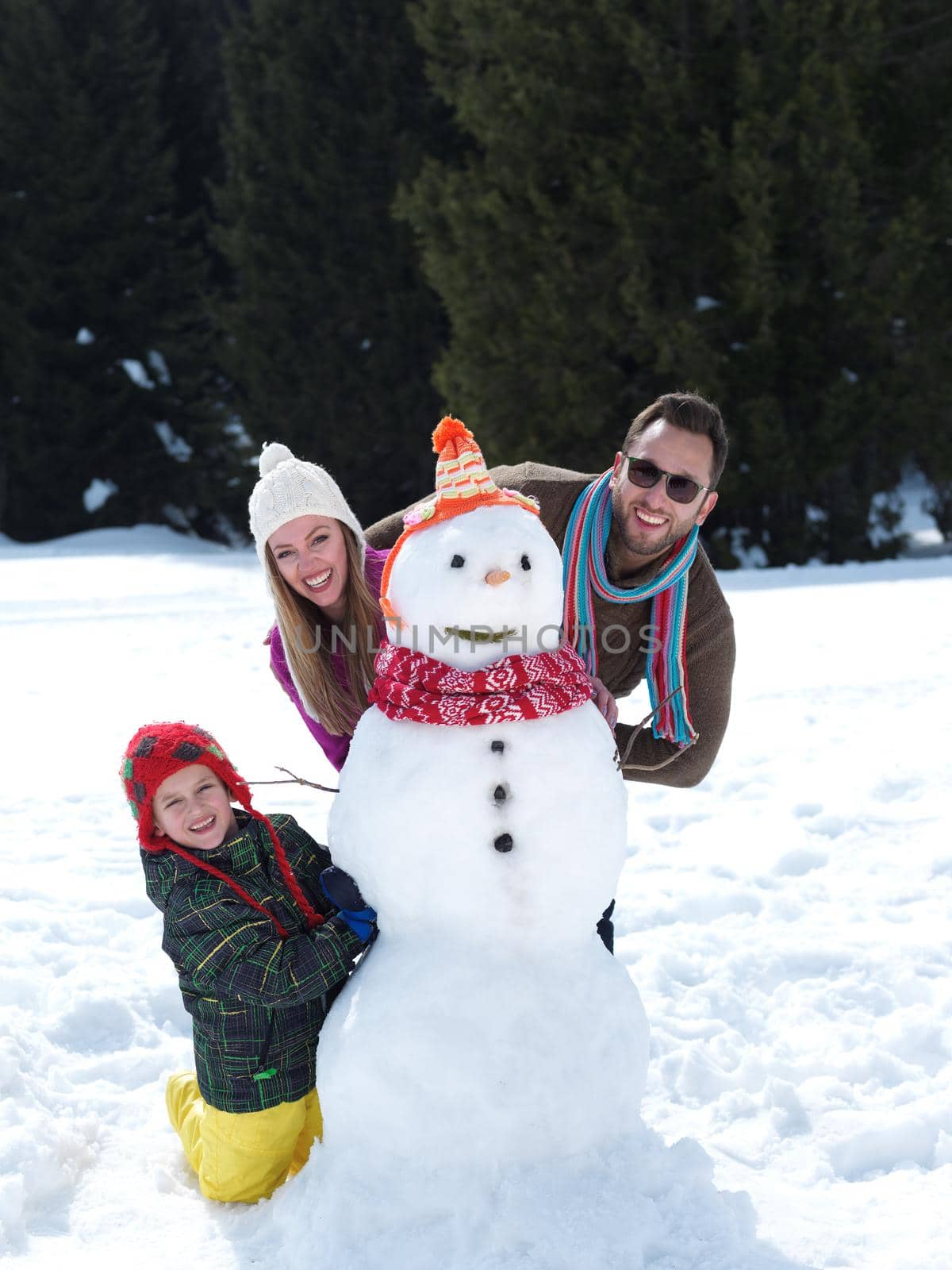 happy young  family playing in fresh snow and making snowman at beautiful sunny winter day outdoor in nature with forest in background