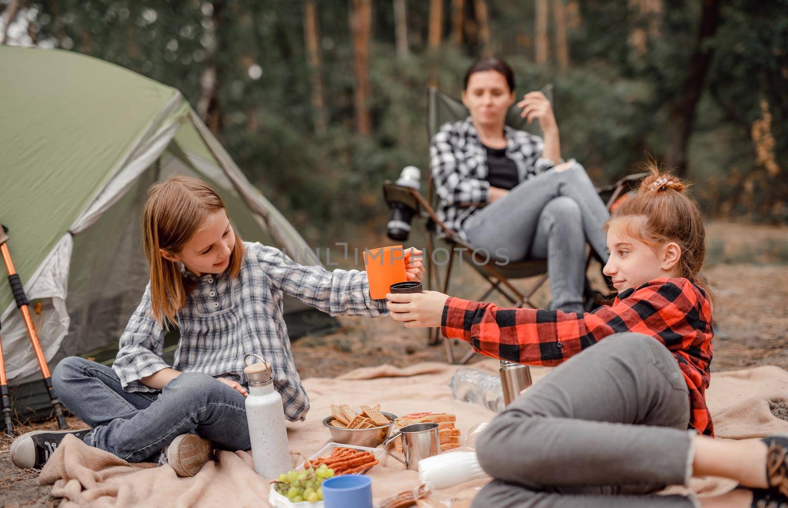 Beautiful girls sisters drinking tea in camping in forest with tent and smiling while their mother sitting and looking at them. Young mother and her daughters resting with picnic in the wood