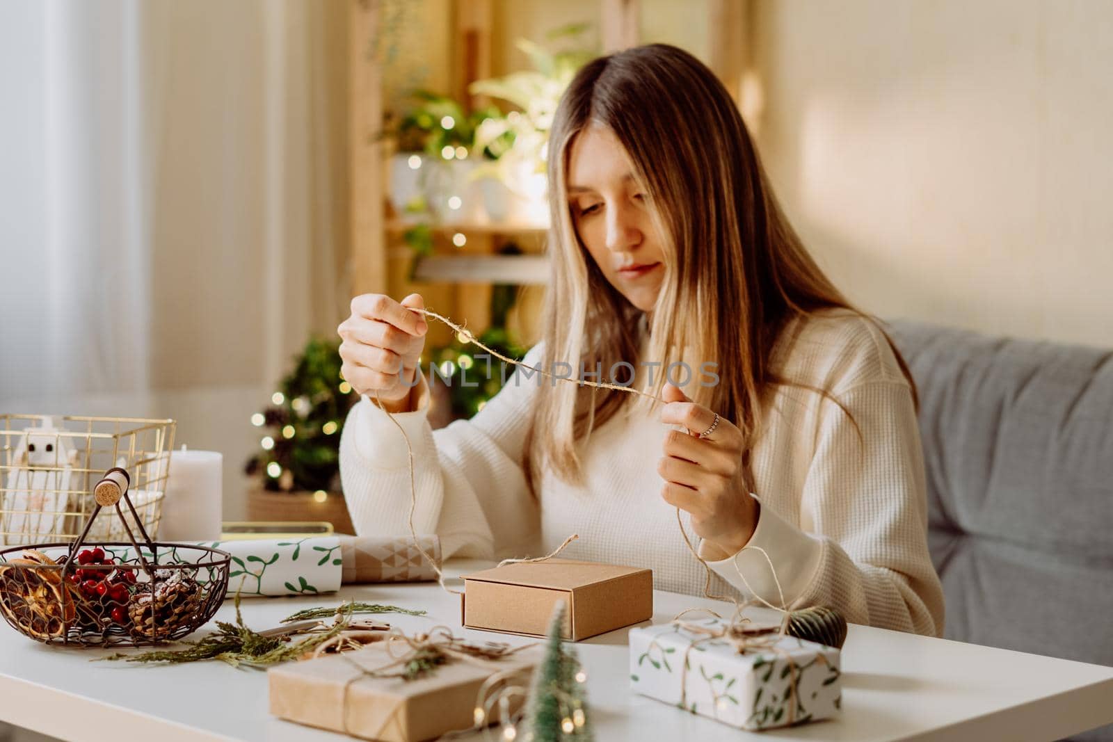 Woman wrapping Christmas eco natural gift boxes at home. Preparing presents on white table with decor elements, lights and items Christmas or New year DIY packing Concept.