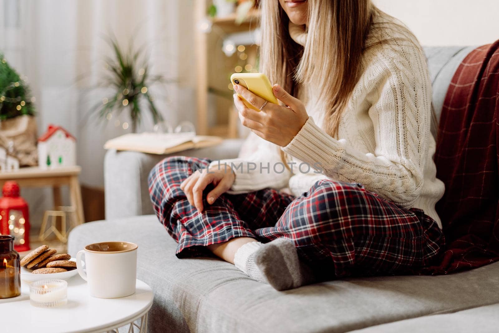 Cozy woman in knitted winter warm socks, sweater and checkered plaid with phone, drinking hot cocoa or coffee in mug, during resting on couch at home. Christmas holidays with candle, decor and cookies