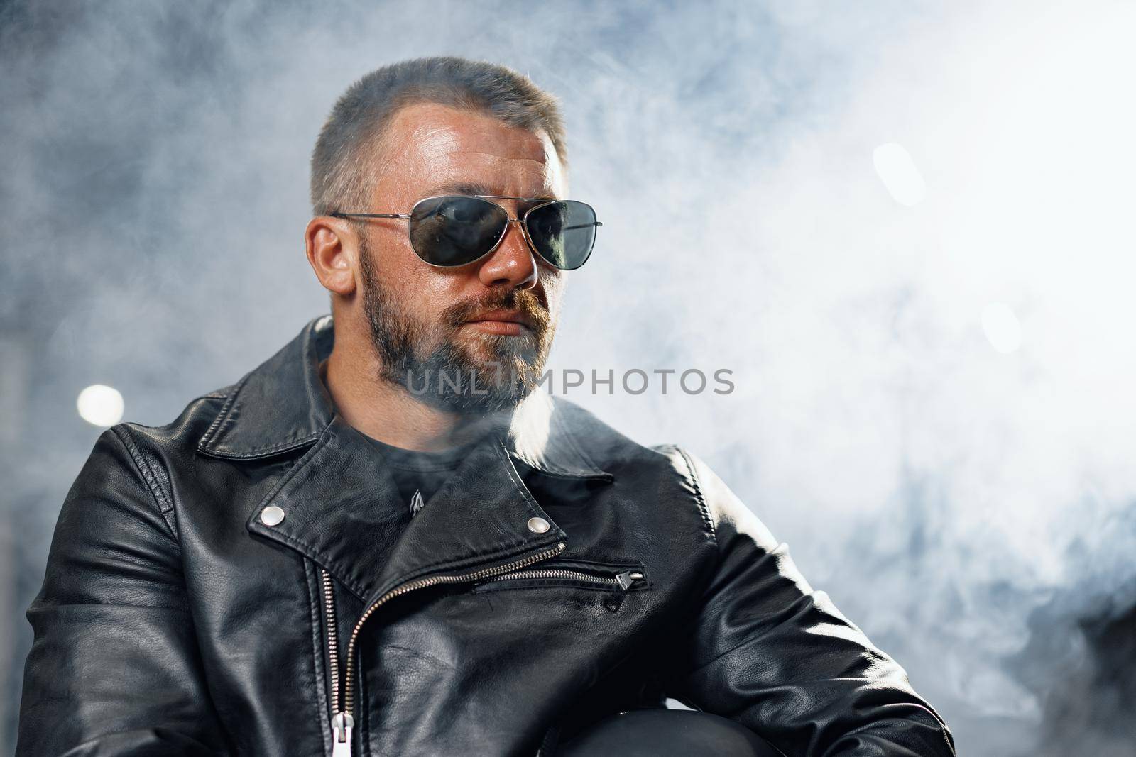 Portrait of bearded man motocyclist in dark sunglasses on dark building background