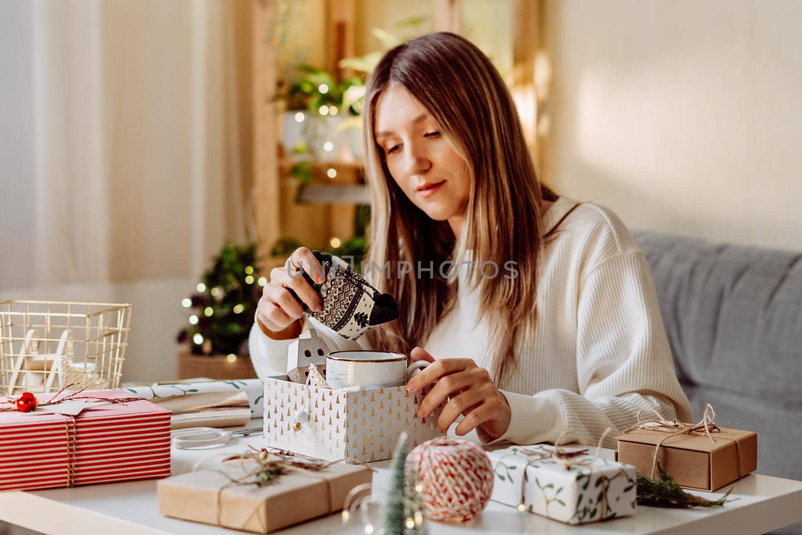 Woman wrapping Christmas eco natural gift boxes at home. Preparing presents on white table with decor elements, lights and items Christmas or New year DIY packing Concept.