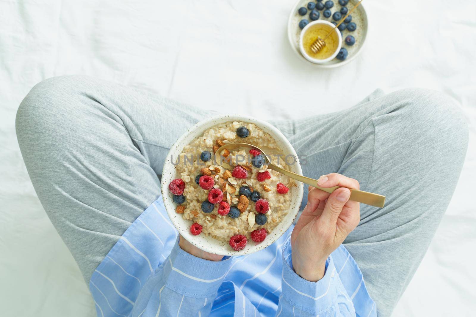 Top view faceless woman holding plate of oatmeal, sitting in bed early morning, quiet home environment. Healthy breakfast
