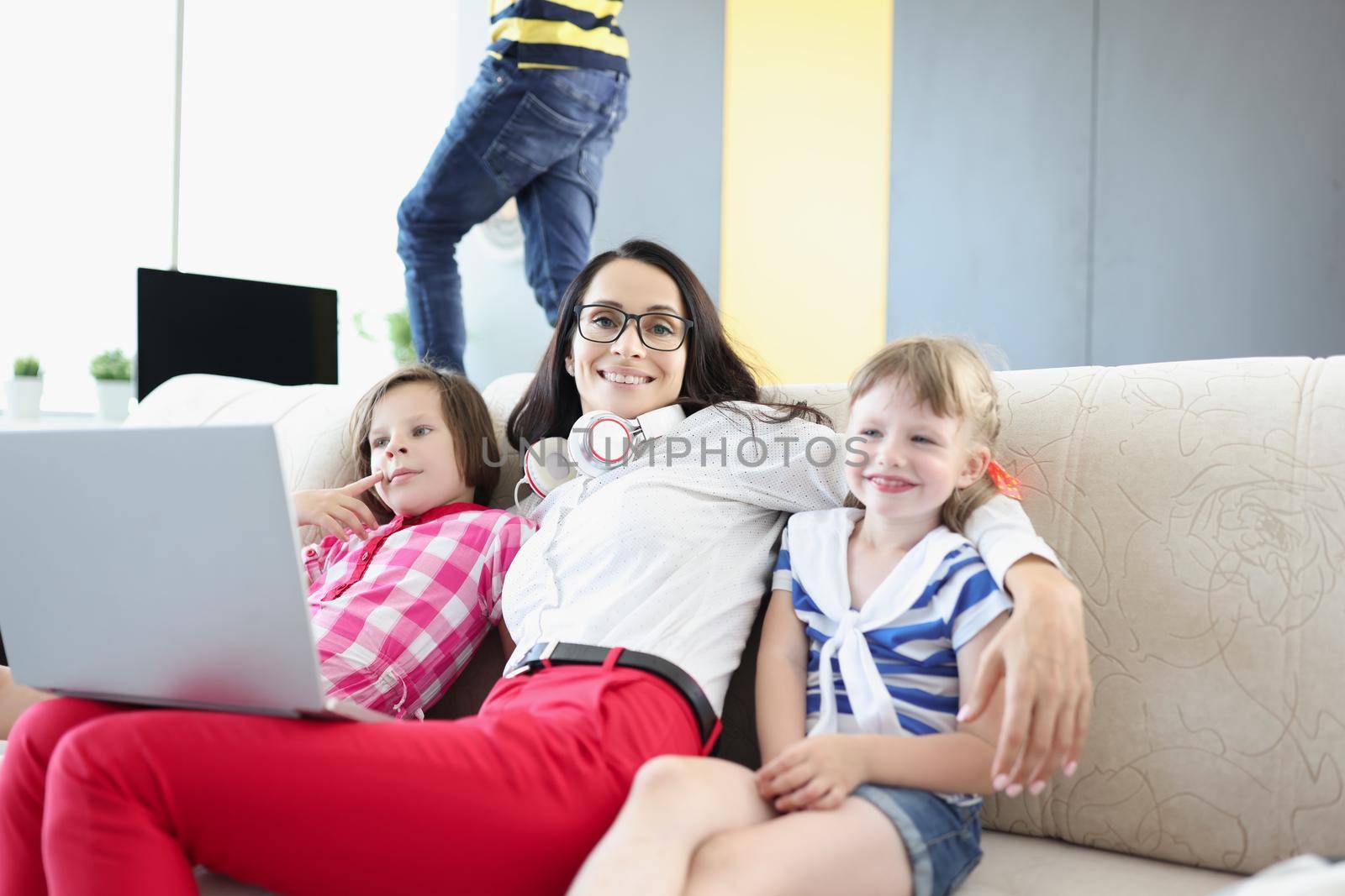 Portrait of happy woman posing with her kids on couch while working on laptop. Mother work from home, earn money for children. Parenthood, weekend concept