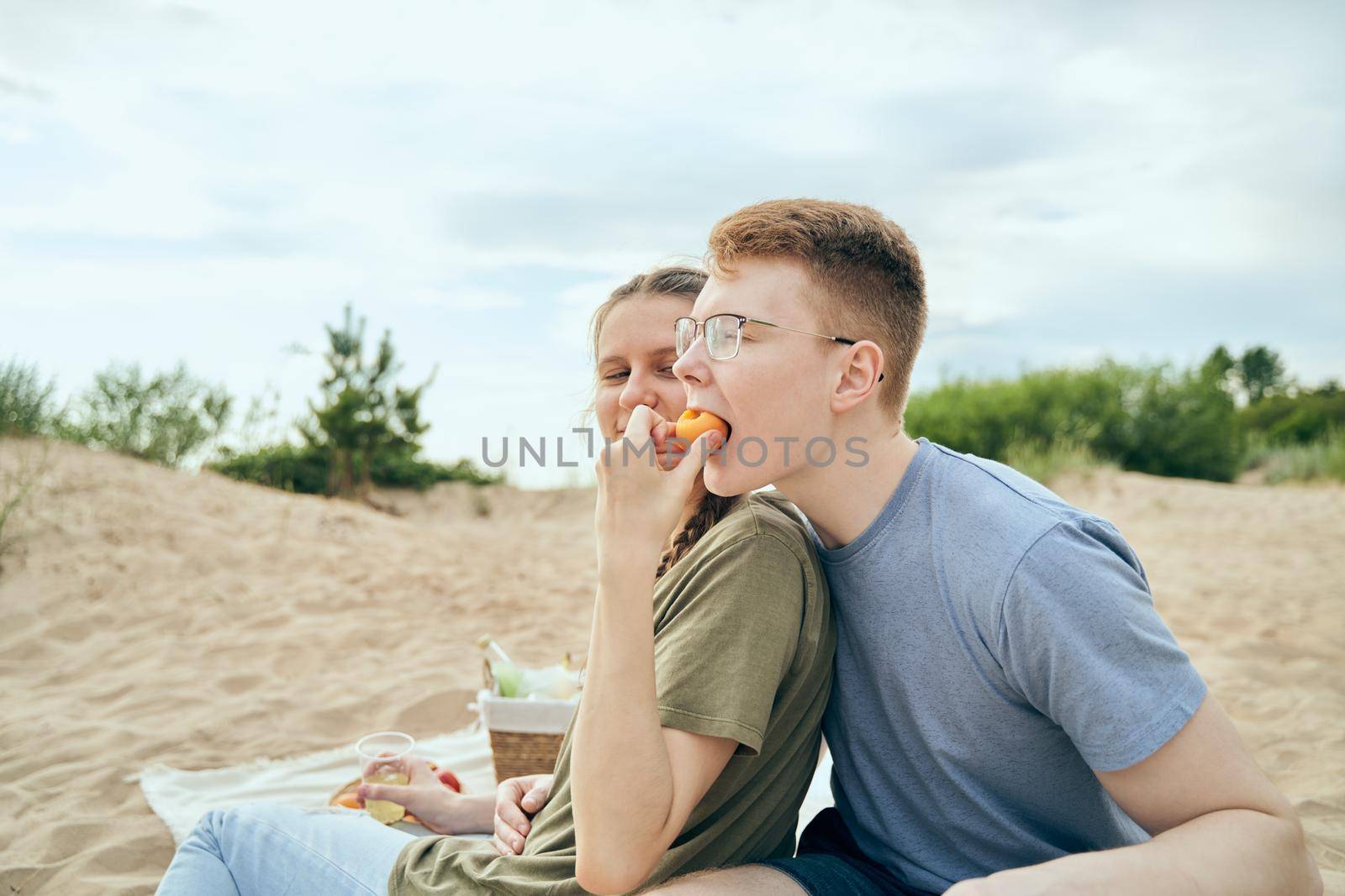Side view of boyfriend and girlfriend feeding each other smiling and holding plastic cups during picnic on seashore background
