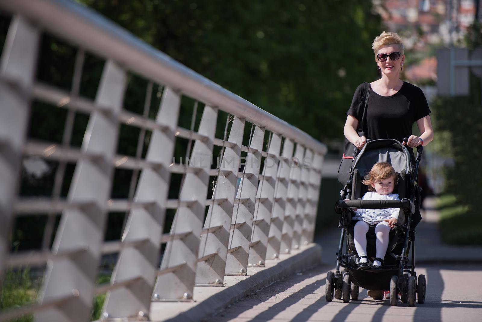 beautiful young mother with blond hair and sunglasses pushed her baby daughter in a stroller on a summer day