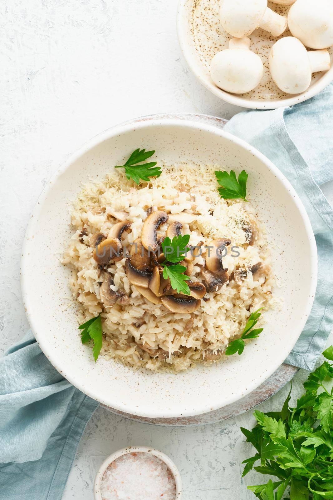 Risotto with mushrooms in plate. Rice porridge with mushrooms and parsley. White table, spoons, mushrooms. Hot dish, italian cuisine, top view, vertical