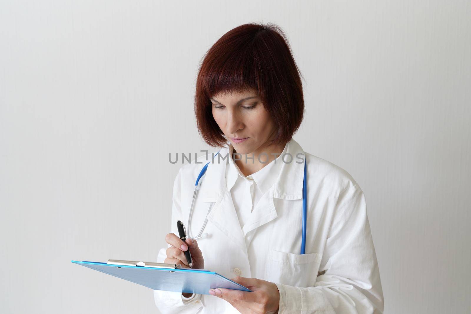 Female adult doctor stands on white background. Beautiful woman, thinking, writing. Clipboard in his hand, stethoscope around his neck .