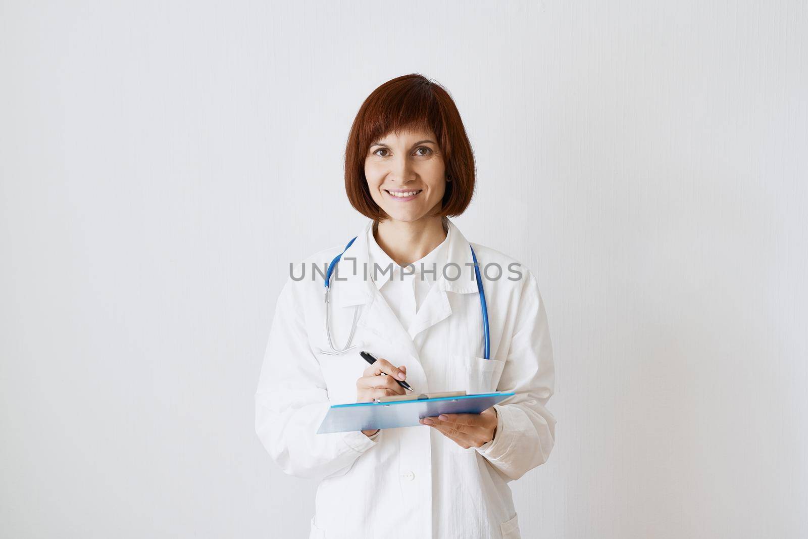 Female adult doctor stands on white background. Beautiful woman, thinking, smiling, writing. Clipboard in his hand, stethoscope around his neck .