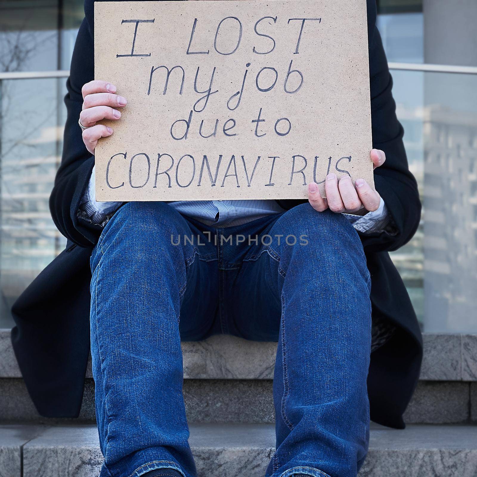 Job loss due to COVID-19 virus pandemic concept. Unrecognizable man holds sign I lost my job because of coronavirus. Male sitting on stairs of building, former place of work in business center