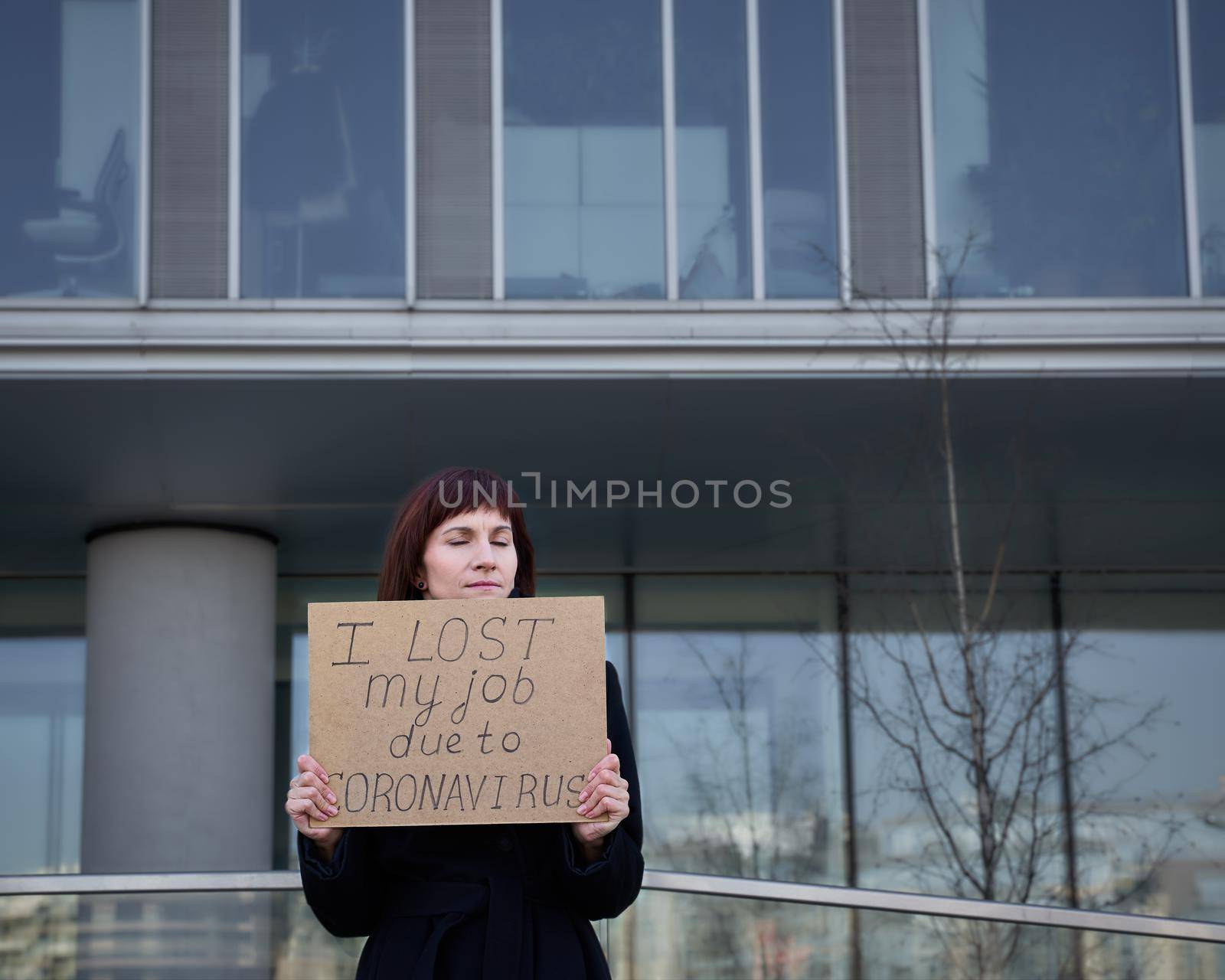Woman holds sign saying I lost my job because of coronavirus. Concept of job loss due to the COVID-19 virus pandemic. Female with closed eyes stands against background of business center