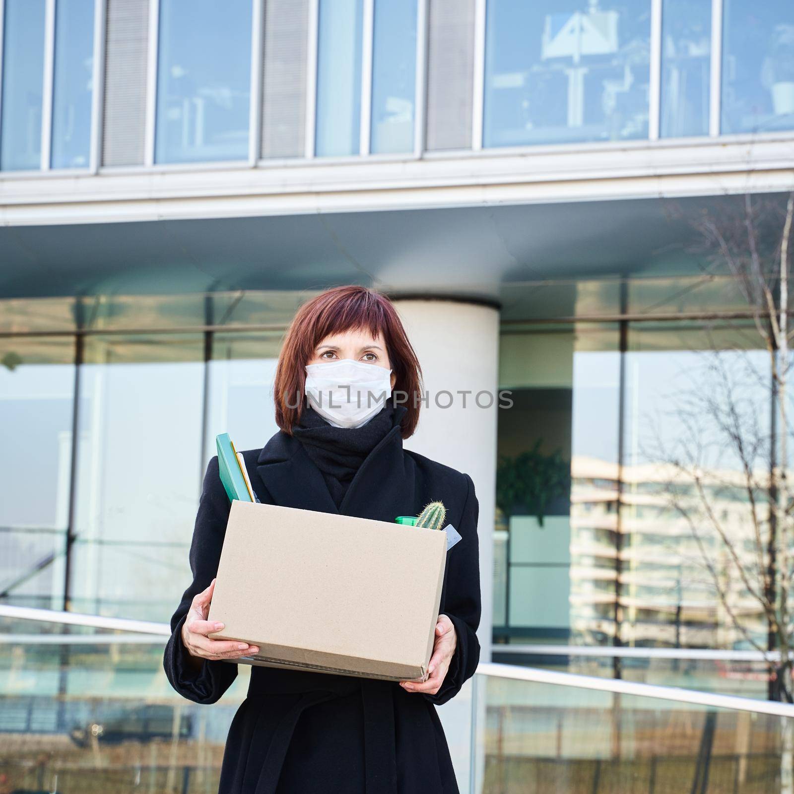Concept of job loss due to the COVID-19 virus pandemic. Woman in mask lost job because of coronavirus. Female with box of his things against background of business center