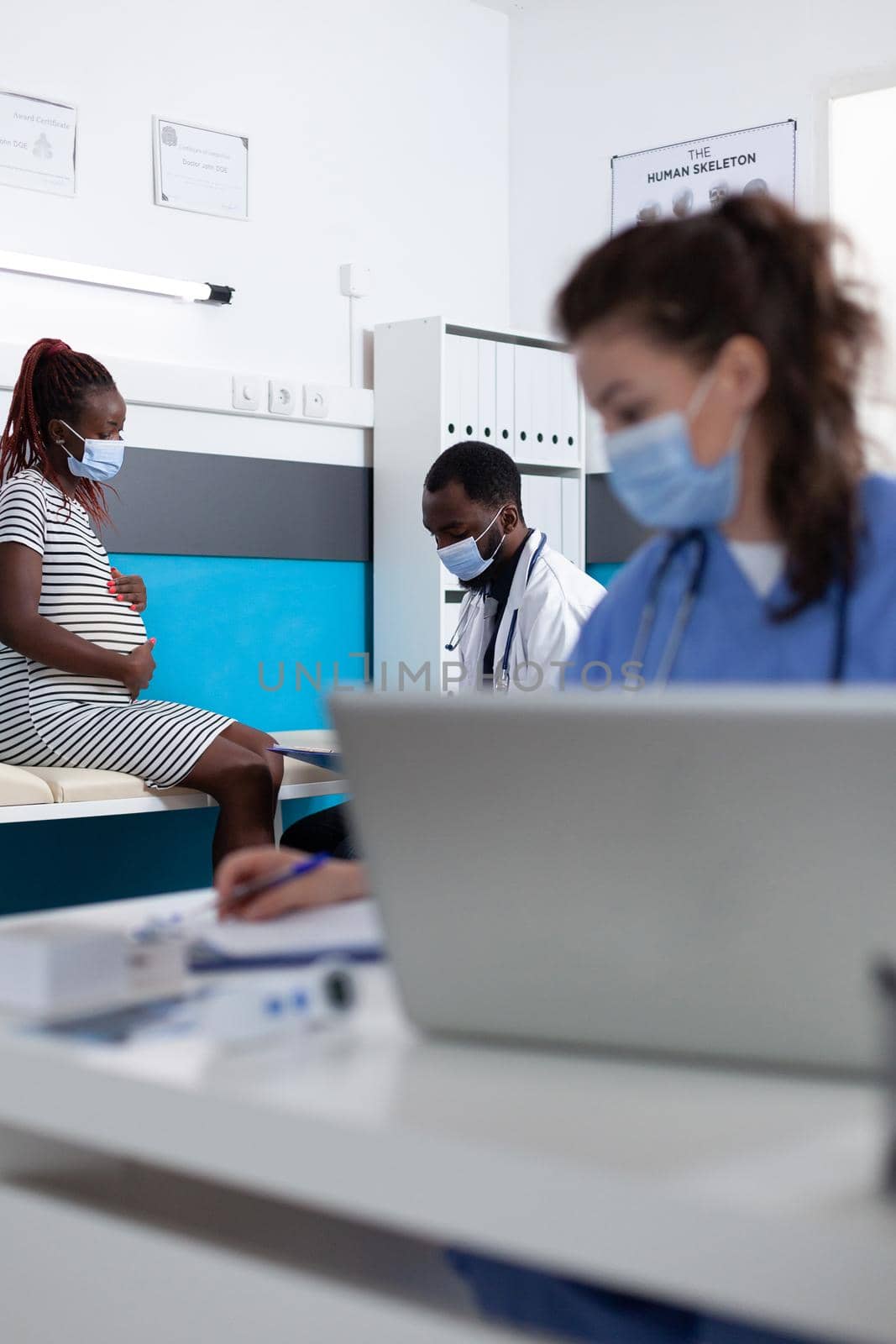 Doctor and patient with face masks talking about pregnancy at medical checkup in office. African american woman expecting child and doing healthcare consultation with practitioner.