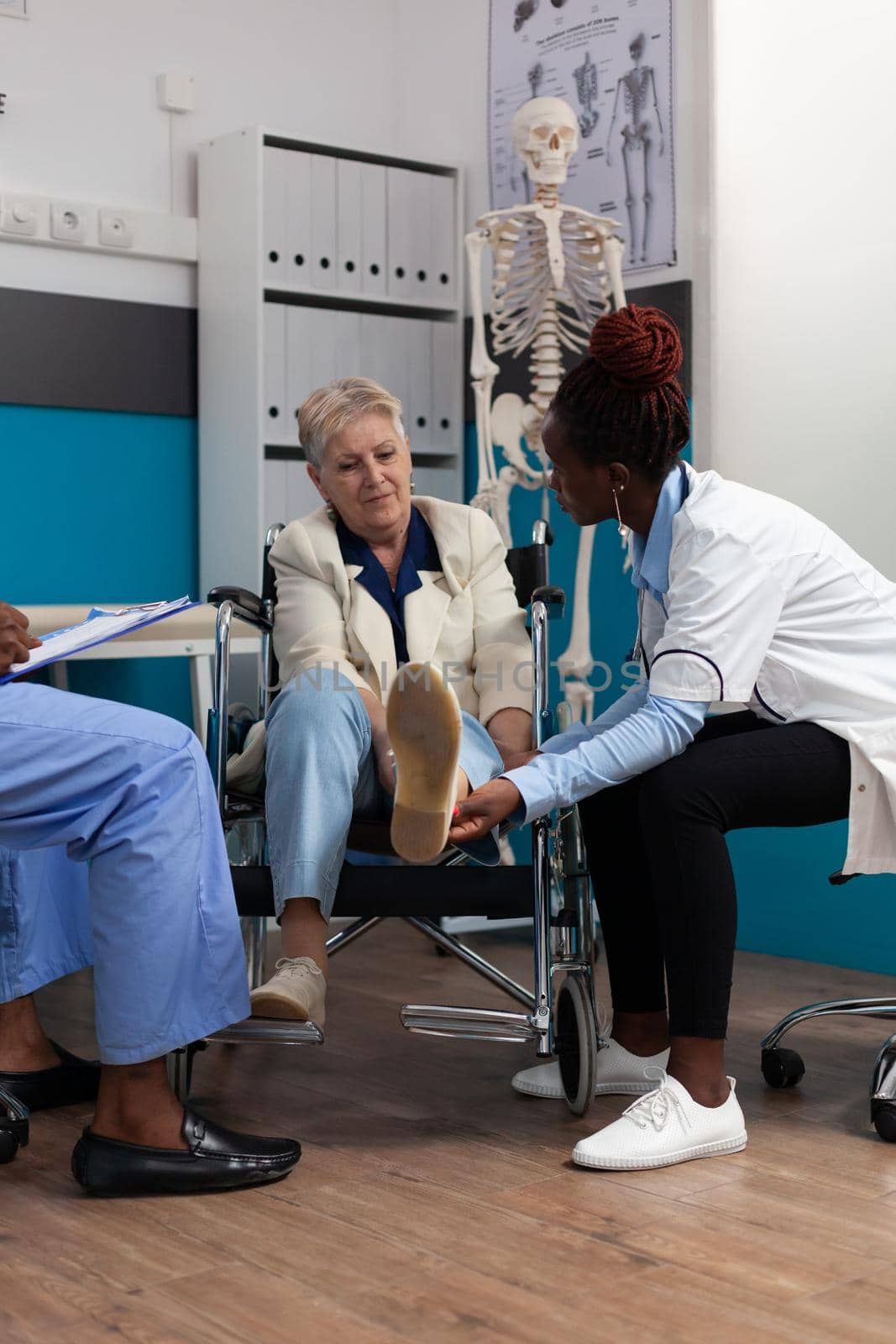 Disabled senior woman geeting knee examined during physiotherapy appointment in hospital office. Therapist doctor massaging injury leg while man nurse writing sickness symptoms. Medical exam concept