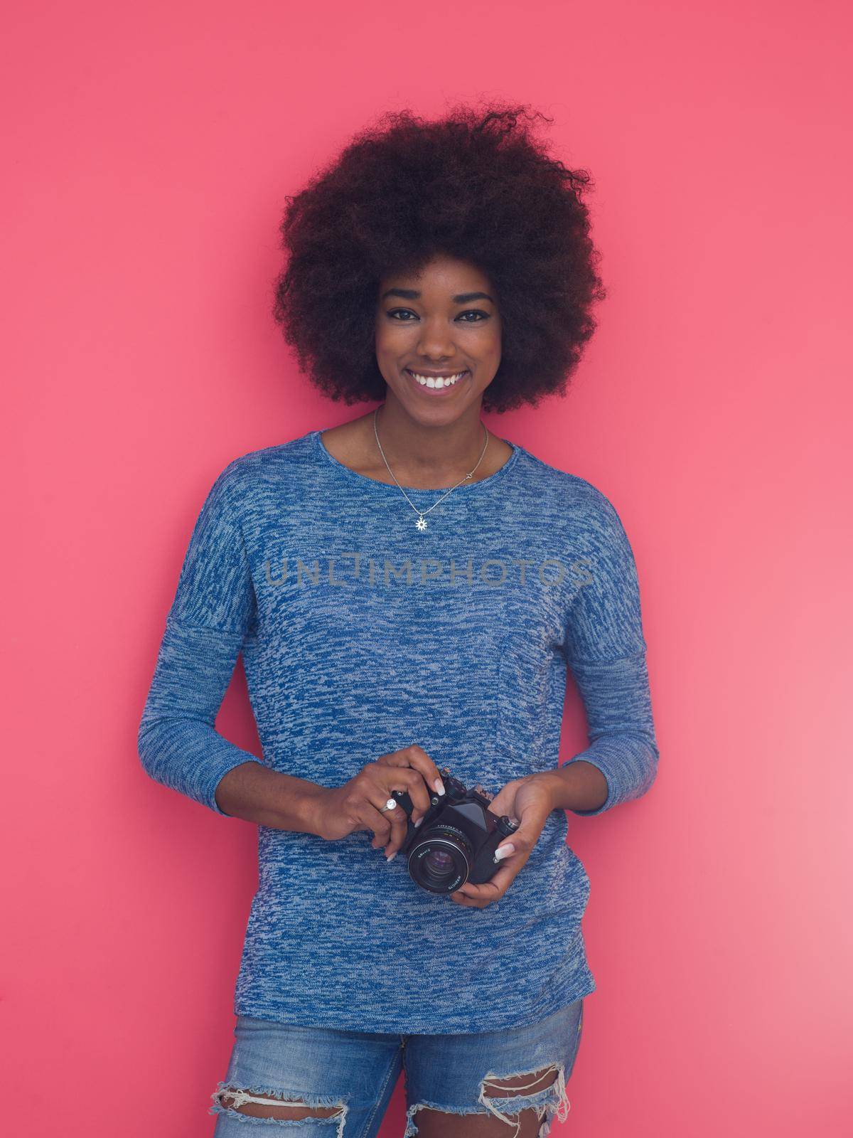 portrait of a smiling pretty african american girl taking photo on a retro camera isolated over pink background