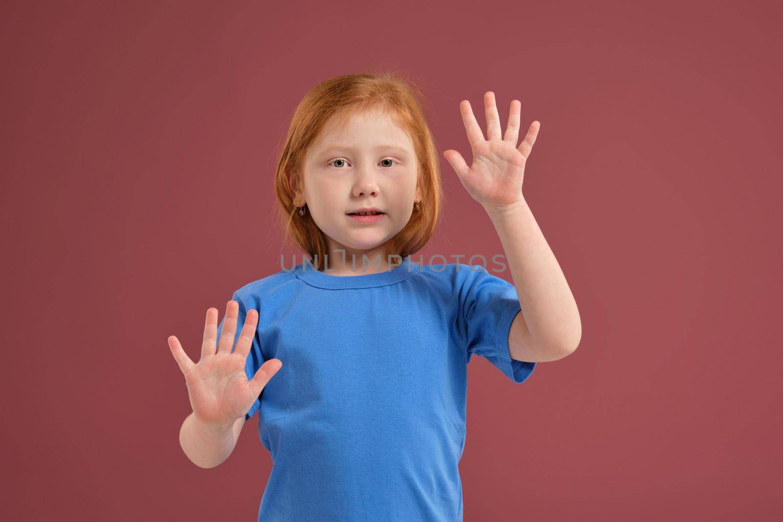 Portrait of cute redhead emotional little girl in blue t-shirt on red background. Beautiful studio portrait of female little caucasian person
