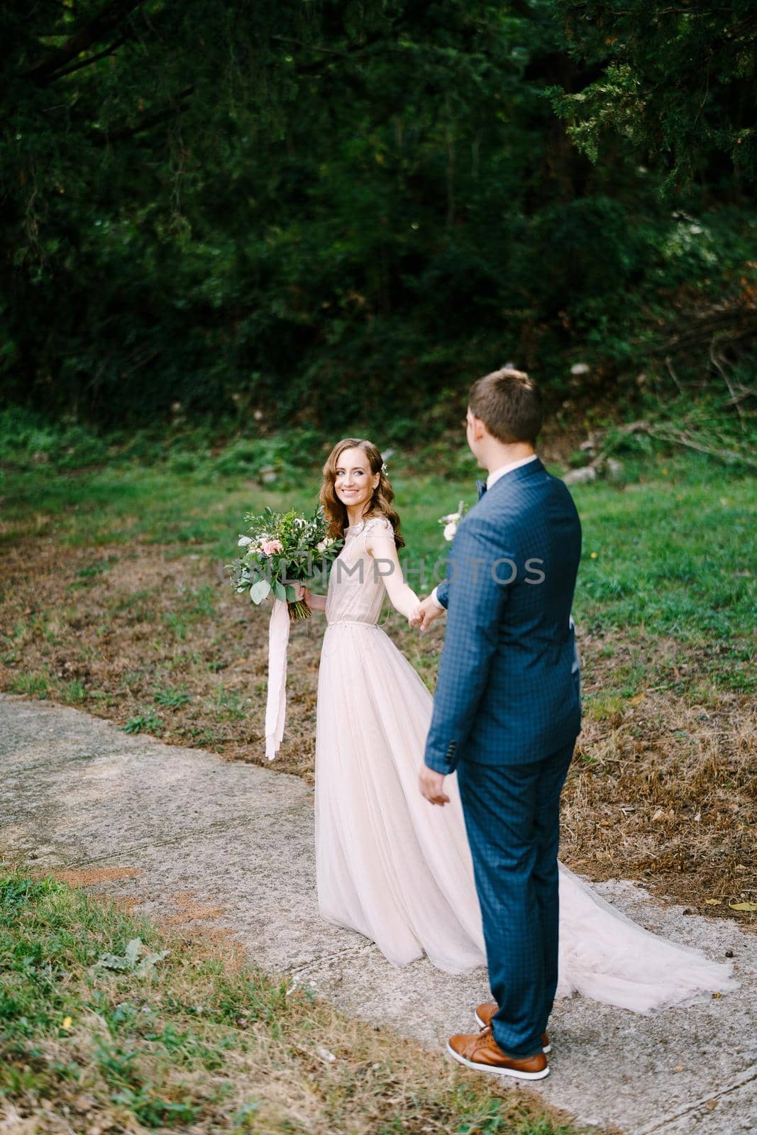 Smiling bride with a bouquet pulls the groom hand along the path in the garden. High quality photo