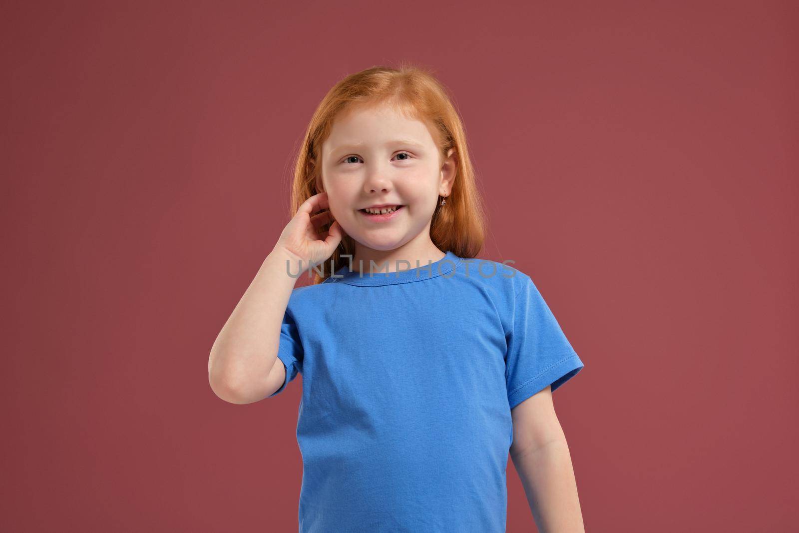 Portrait of cute redhead emotional little girl in blue t-shirt on red background. Beautiful studio portrait of female little caucasian person. SHe looks very happy