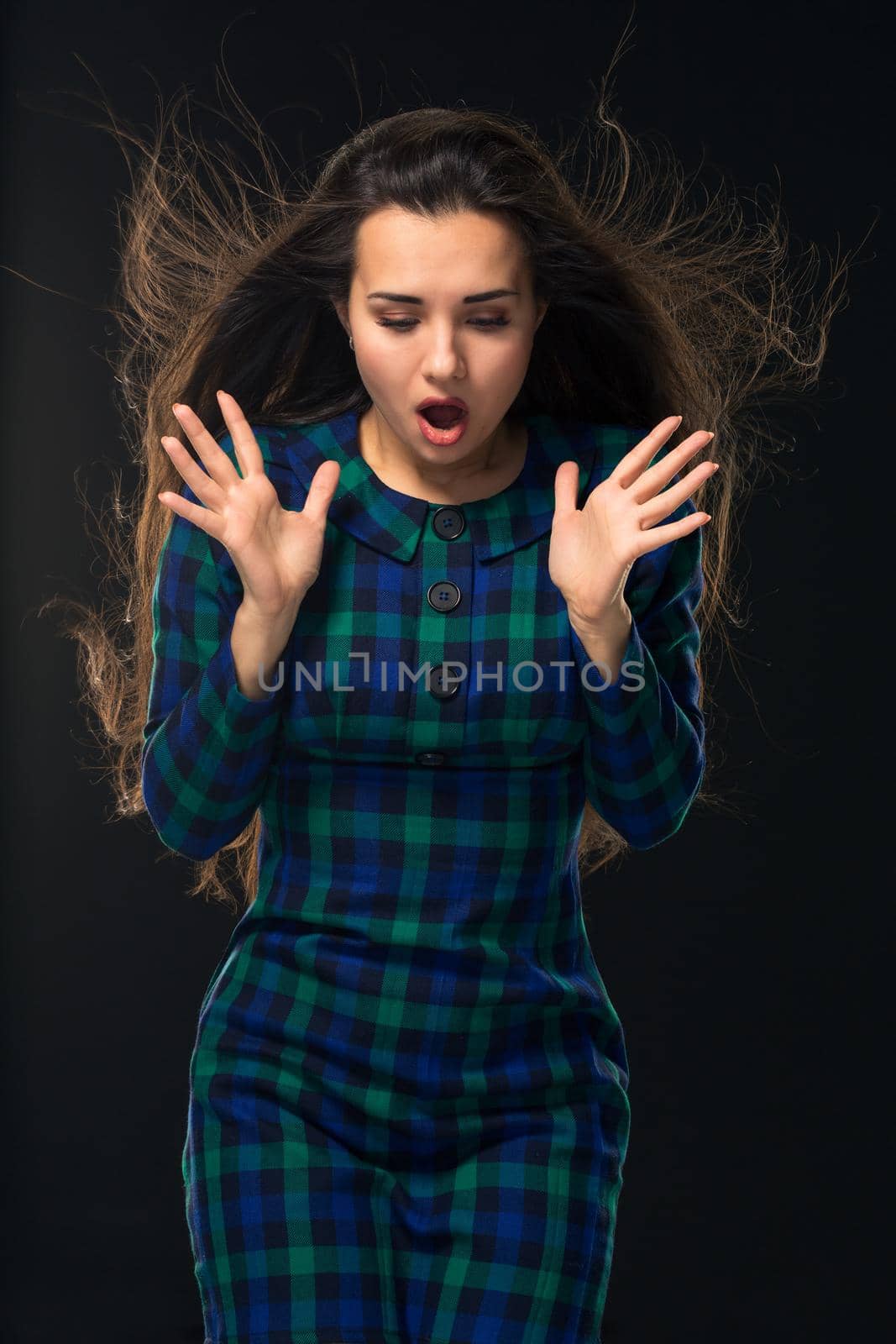 Portrait of cheerful attractive modern woman with long hair, expressing emotions while standing over black background. Young woman in blue checkered dress