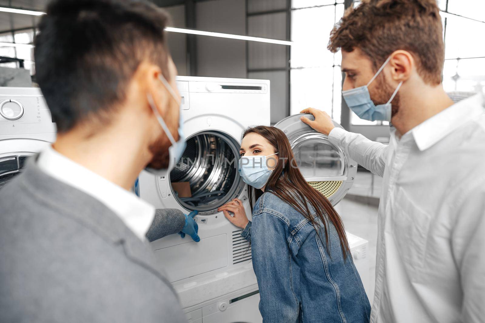 Salesman in hypermarket wearing medical mask demonstrates his clients a new washing machine, close up