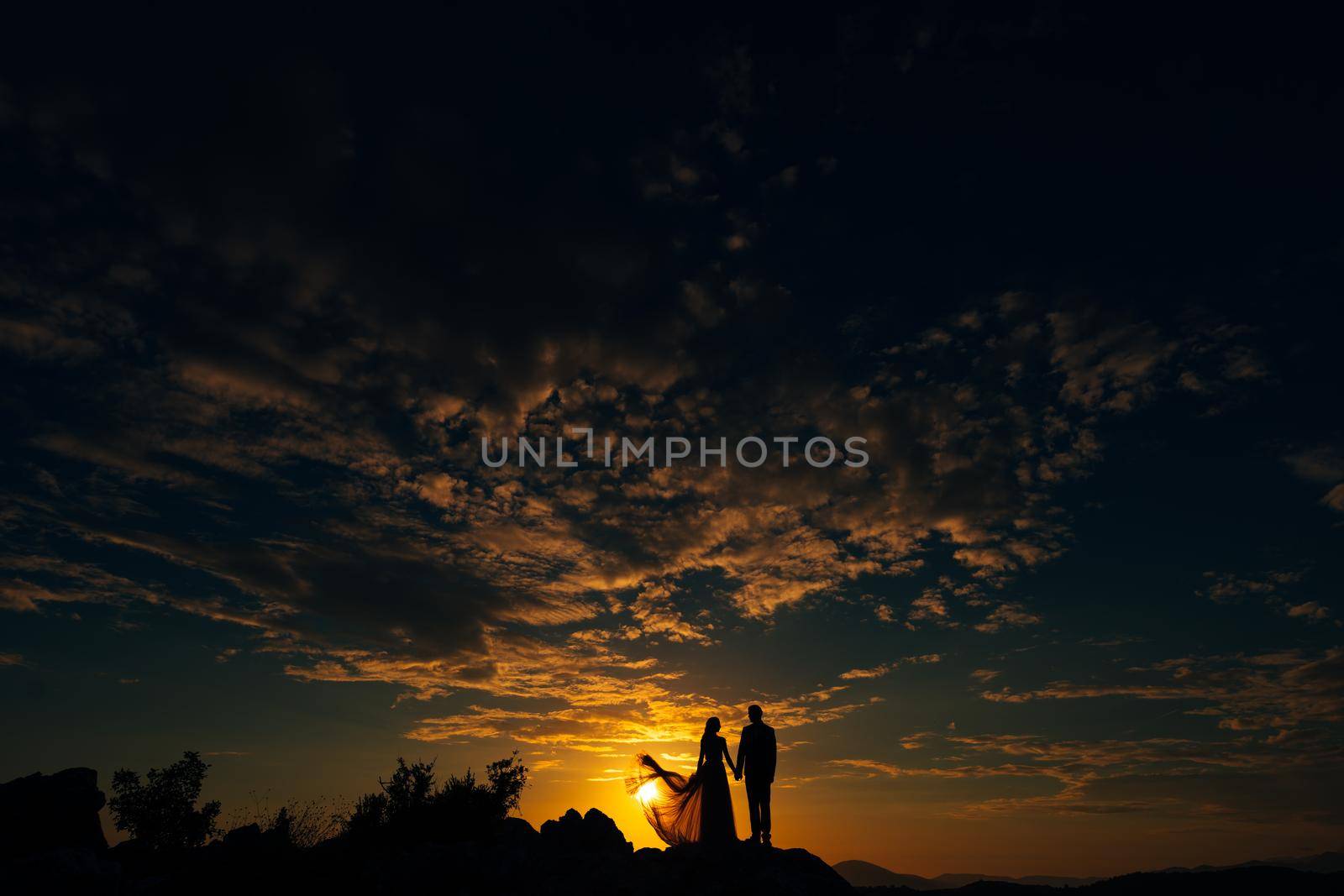 Groom and bride stand on the mountain against the background of the sunset sky by Nadtochiy