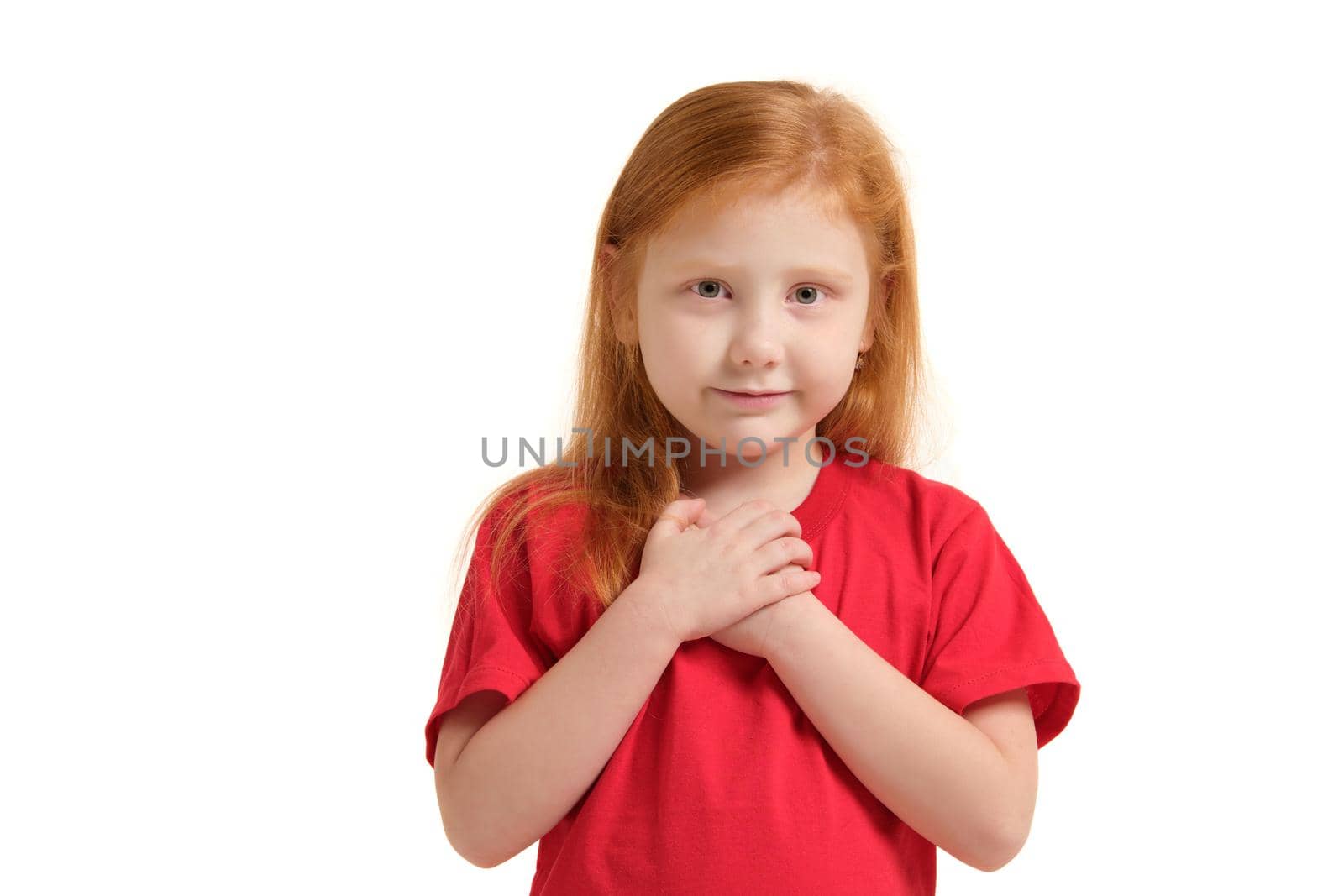 Portrait of cheerful caucasian little girl with red hair posing in studio with looking to the camera. Girl dressed in red T-shirt keeping palms pressed together, having begging look.