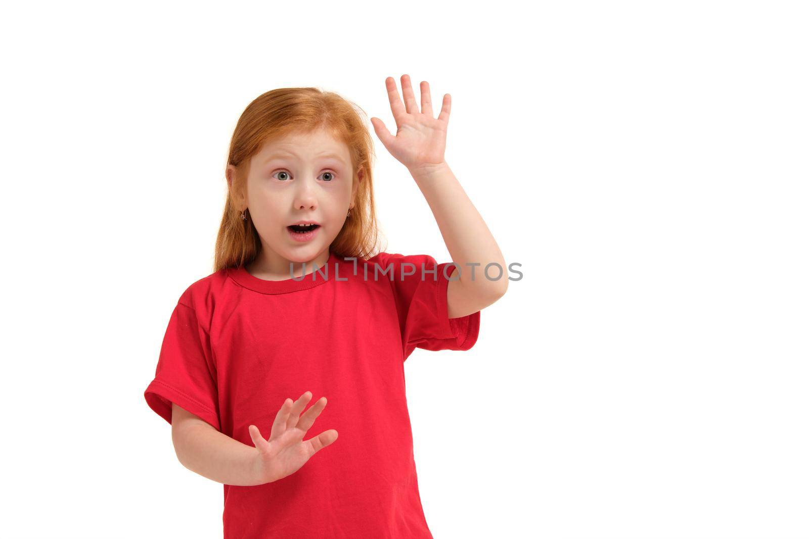 Portrait of cute redhead emotional little girl in red t-shirt isolated on a white, surprised and touching invisible fake wall. Beautiful studio portrait of female little caucasian person
