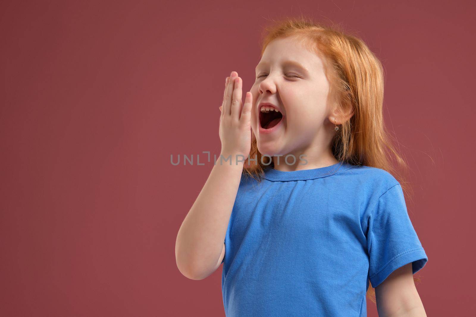 Portrait of cute redhead emotional little girl in blue t-shirt on red background. She is tired and wants to sleep, yawning and stretching