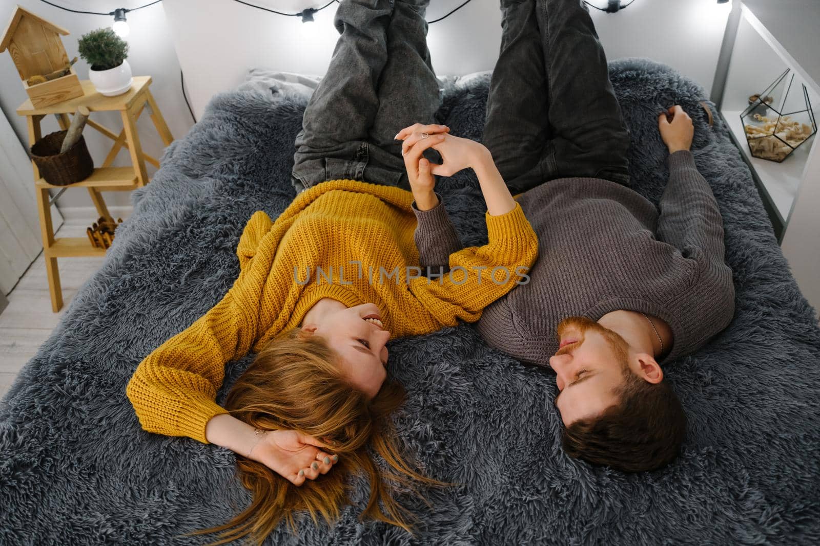 A couple of lovers are lying on a bed with a gray fluffy blanket. Garland at the head of the bed. A girl and a guy hold hands and look at each other.