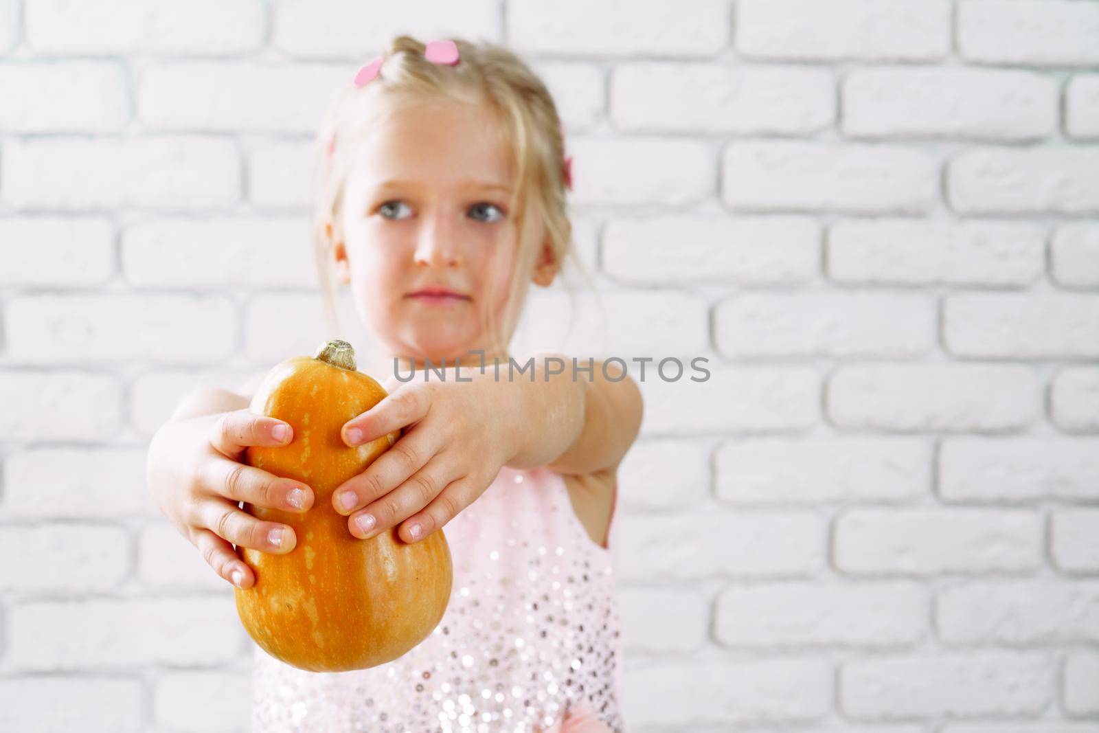 Cute little girl in pink dress holding pumpkin in her hands against white brick wall