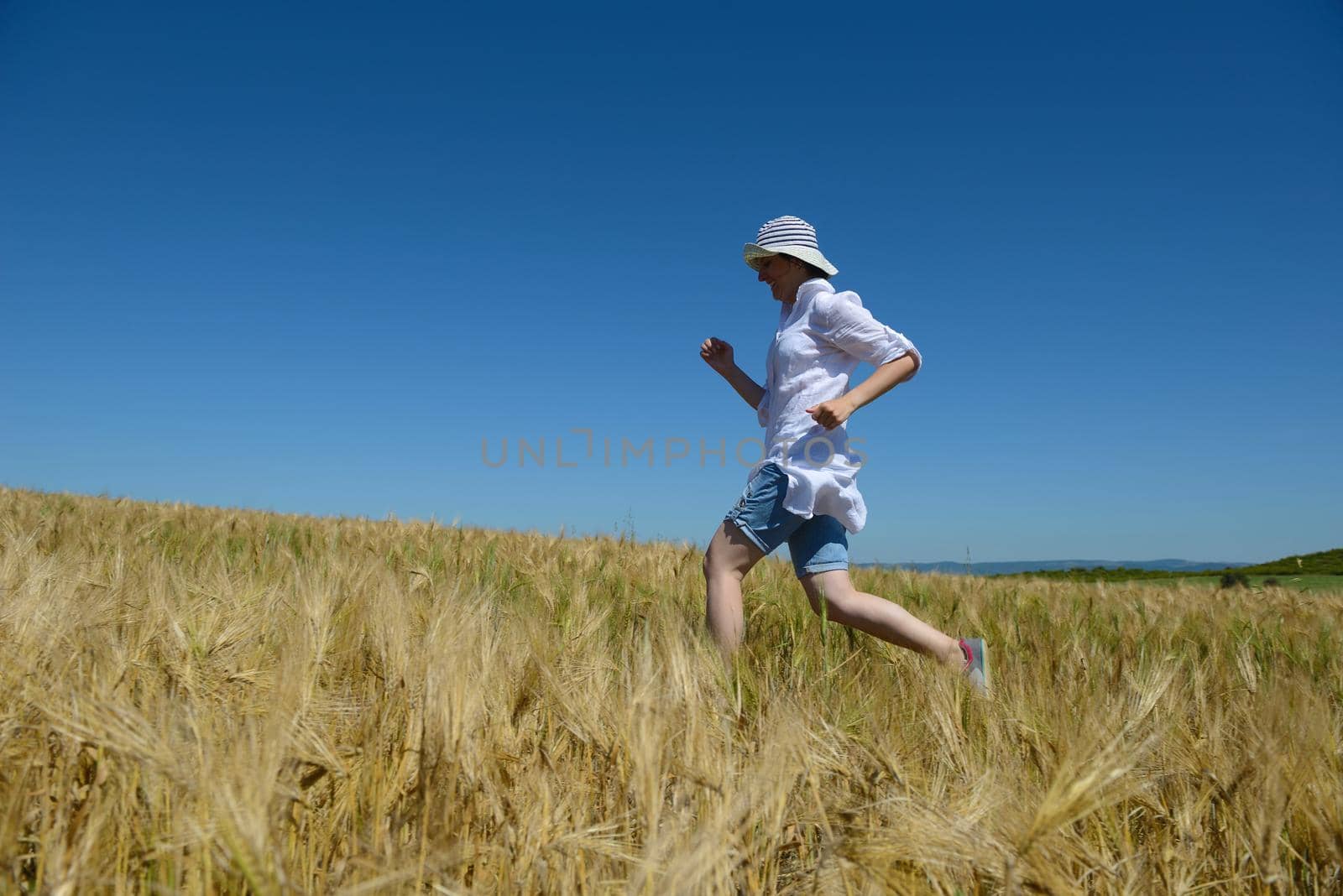 Young woman standing jumping and running  on a wheat field with blue sky in  background at summer day representing healthy life and agriculture concept