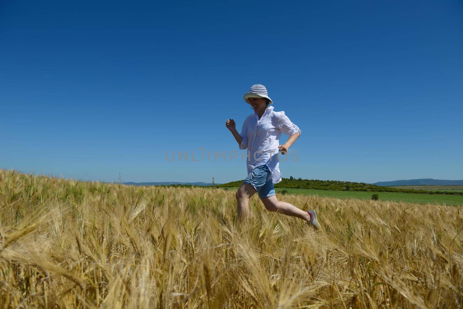 Young woman standing jumping and running  on a wheat field with blue sky in  background at summer day representing healthy life and agriculture concept