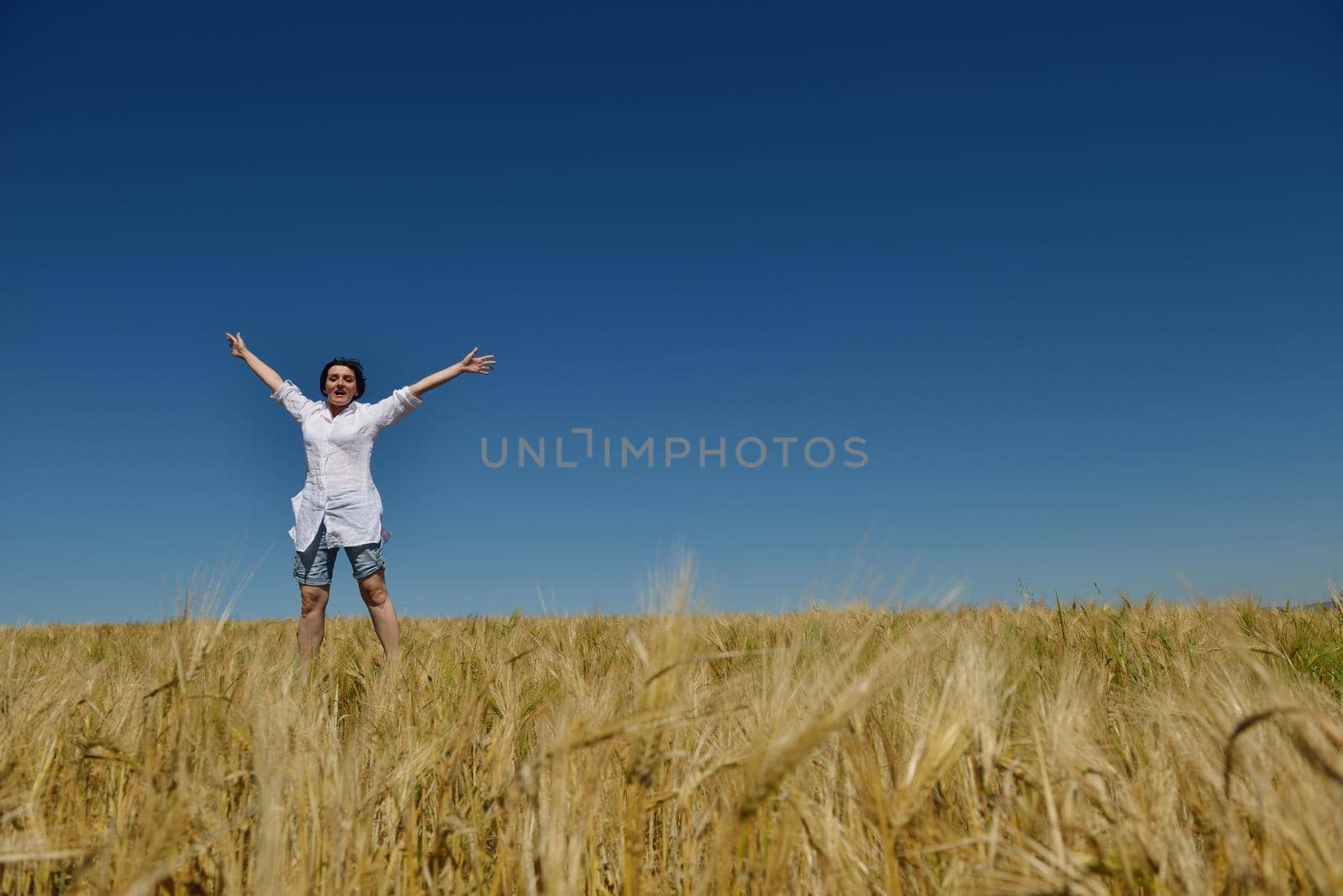 Young woman standing jumping and running  on a wheat field with blue sky in  background at summer day representing healthy life and agriculture concept