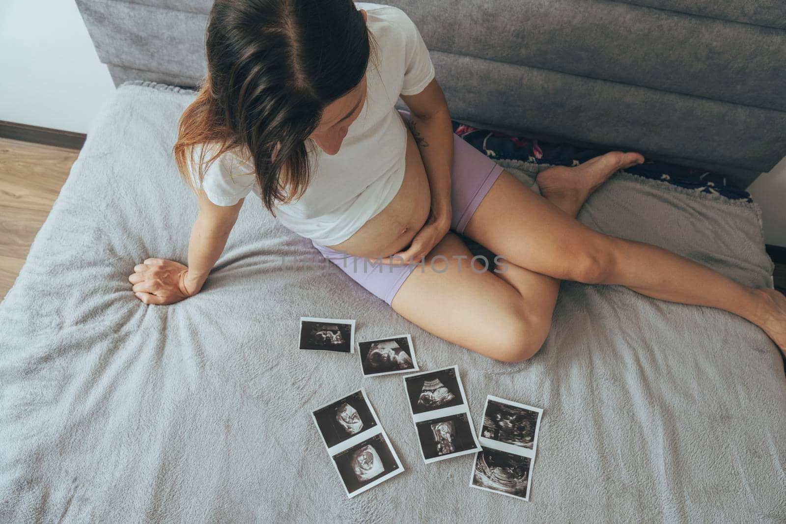 Young pregnant woman, future mother, sitting on comfortable bed and looking at ultrasound x ray photos of her baby in tummy, view from the top.