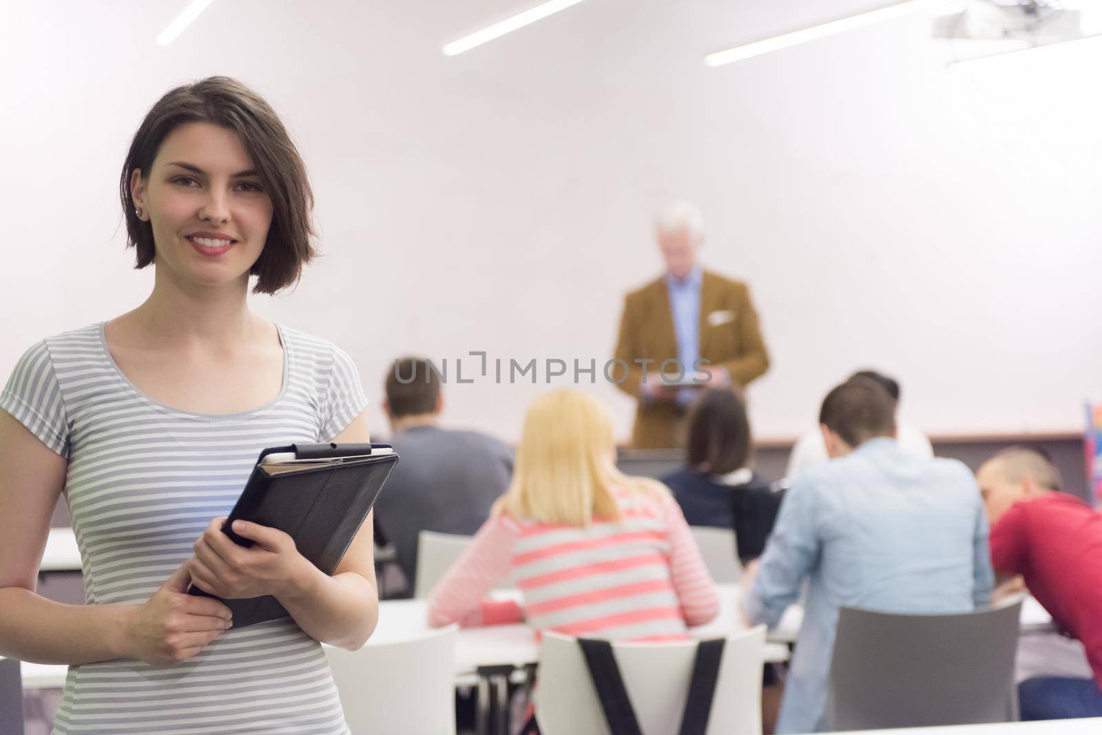 portrait of happy female student holding tablet while teacher teaching students in school classroom