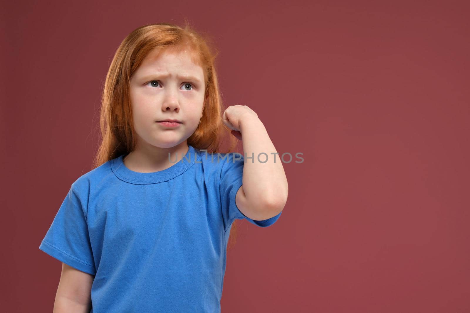close-up portrait of cute redhead emotional little girl in blue t-shirt on red background. She is about to get nervous, angry with someone