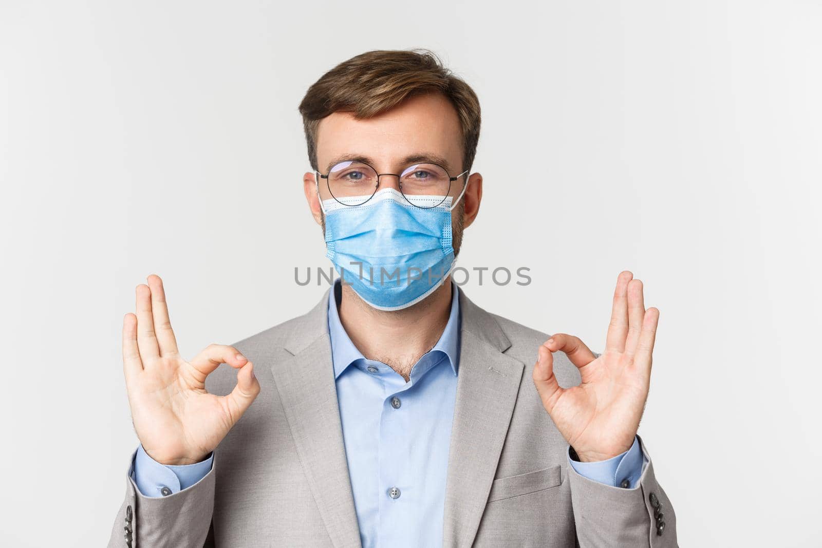 Concept of work, covid-19 and social distancing. Close-up of businessman in grey suit and medical mask, showing okay signs in approval, standing over white background by Benzoix