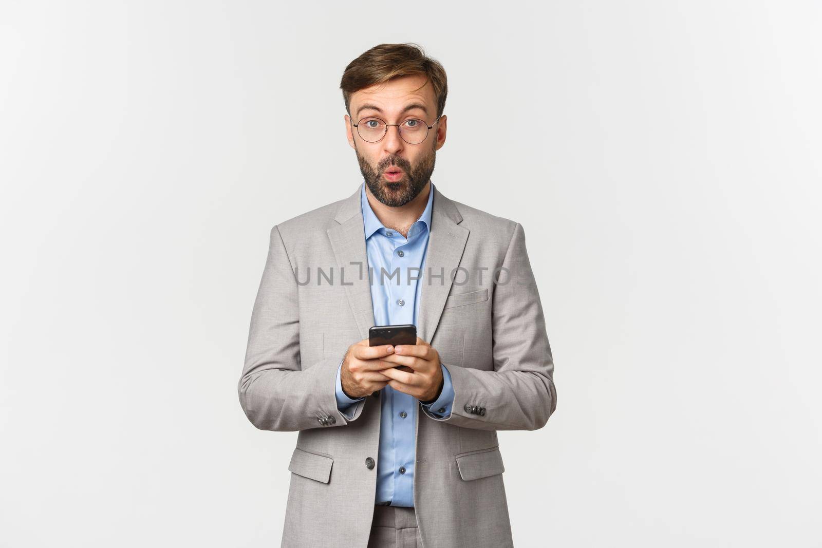 Portrait of handsome businessman with beard, wearing grey suit and glasses, looking amused after reading something interesting on mobile phone, standing over white background.