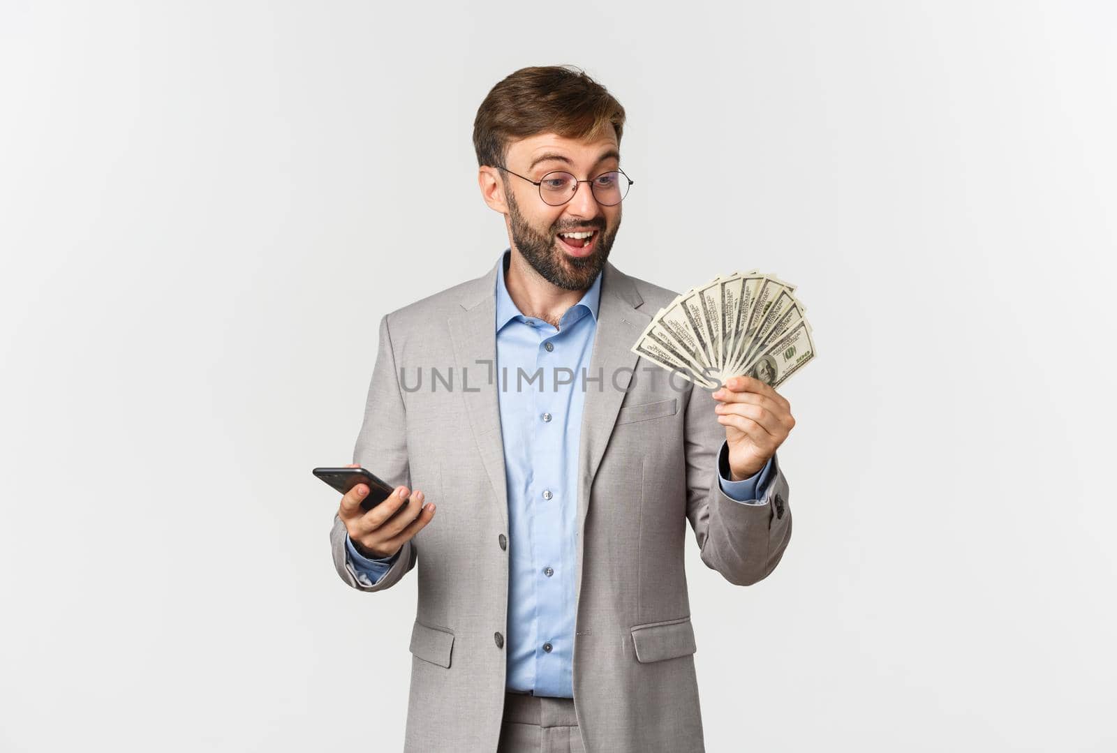 Image of rich successful businessman with beard, looking at money and holding mobile phone, smiling excited, standing over white background.
