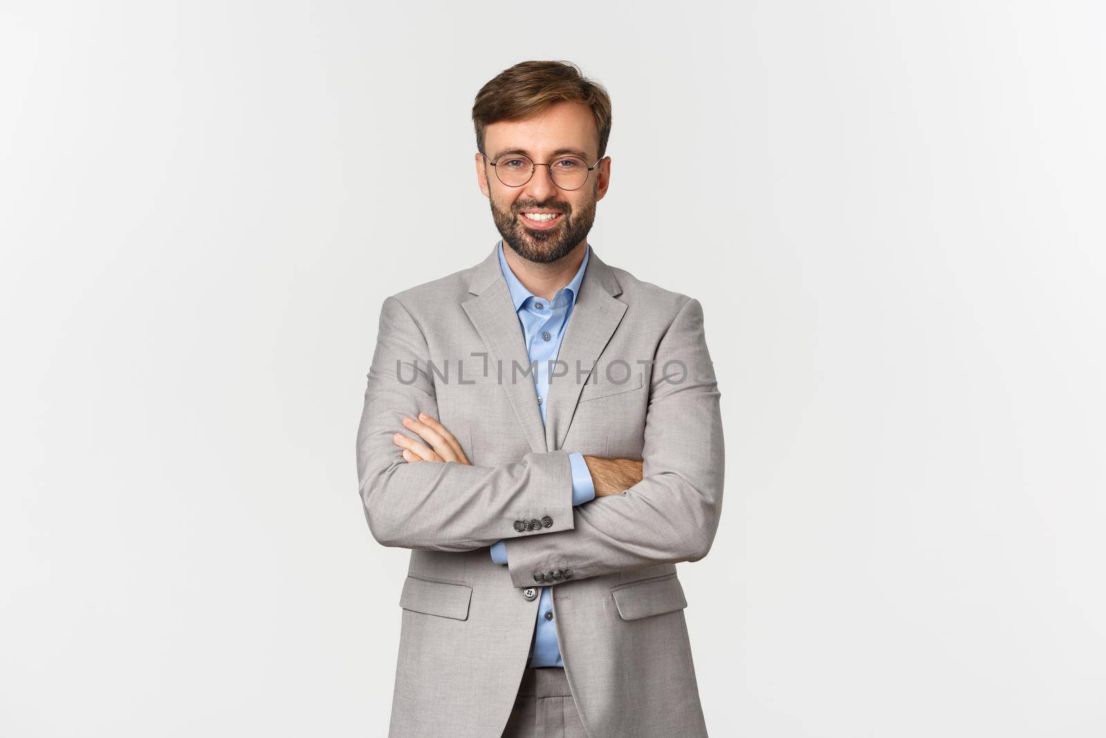 Image of successful and confident businessman with beard, wearing grey suit and glasses, cross arms on chest and smiling satisfied, standing over white background.