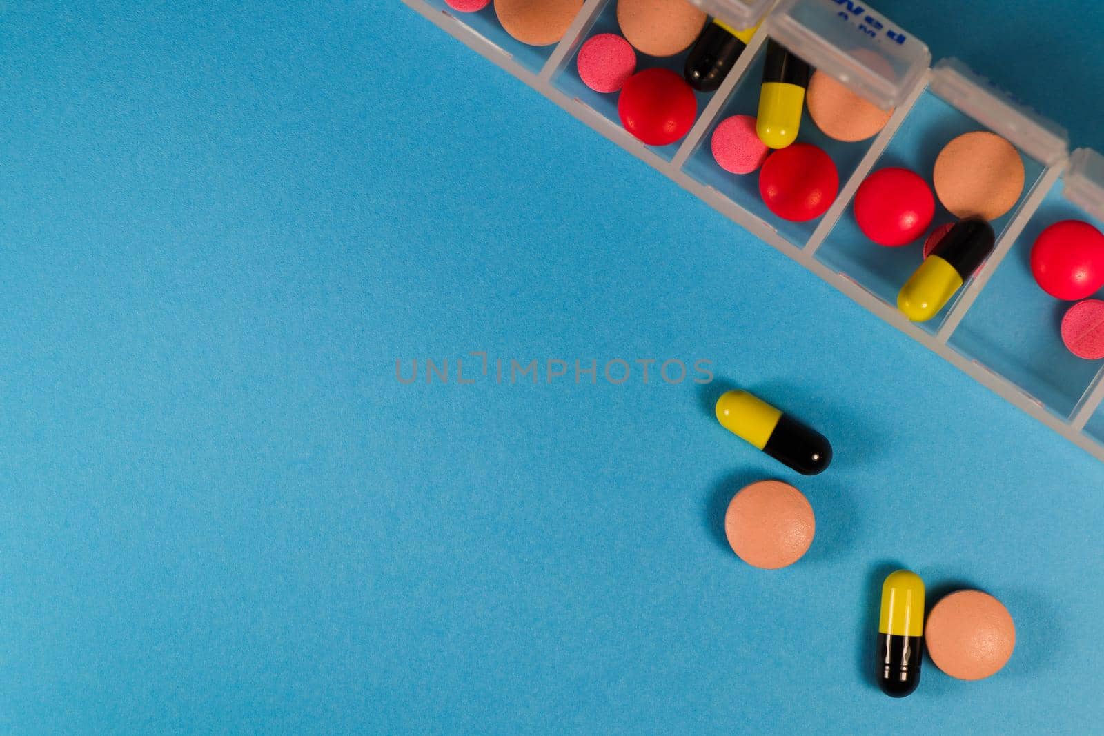 Variety of pills and capsules with a daily prescription drug sorting container on blue background flat-lay with copy space