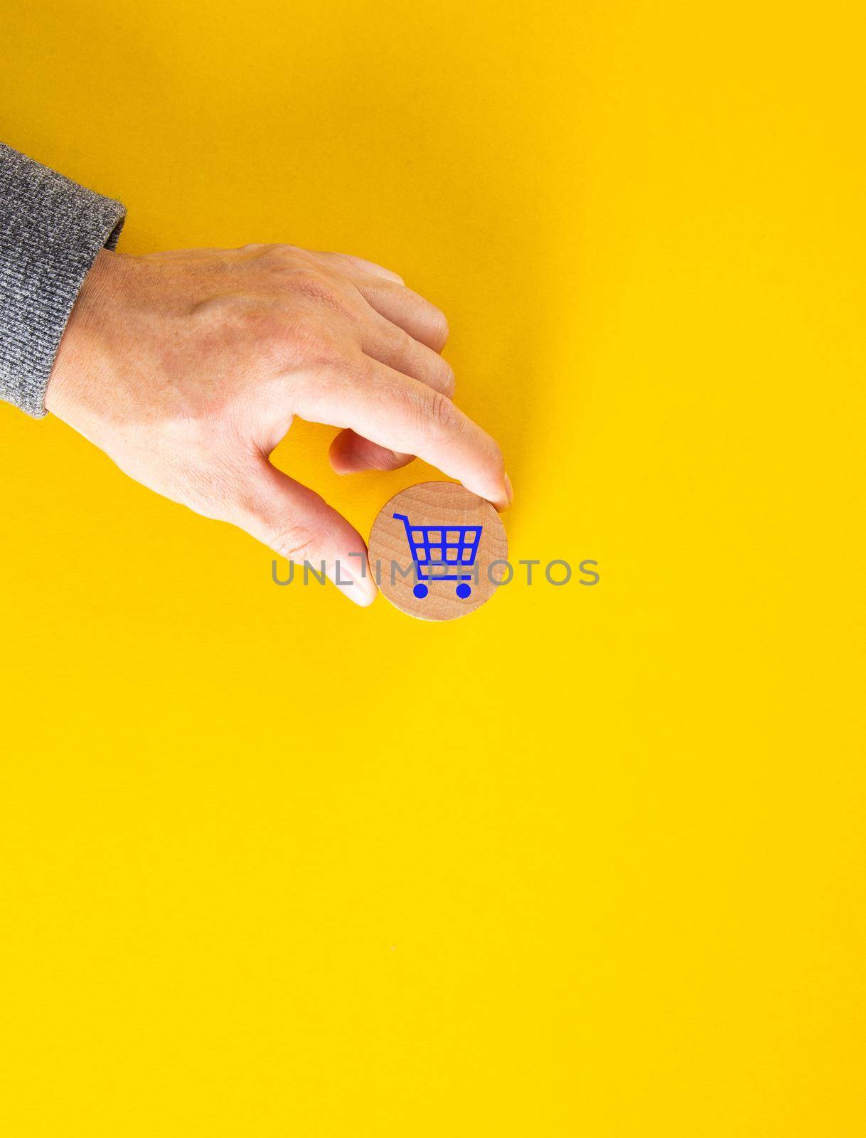 Male hand placing a blue shopping cart icon on yellow background. Shopping or online e-commerce concept.