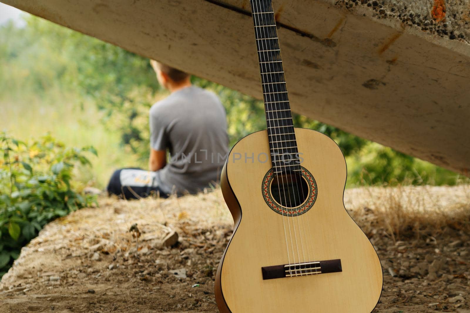 Acoustic guitar, six strings. Boy is sitting on raised platform. Summer day.