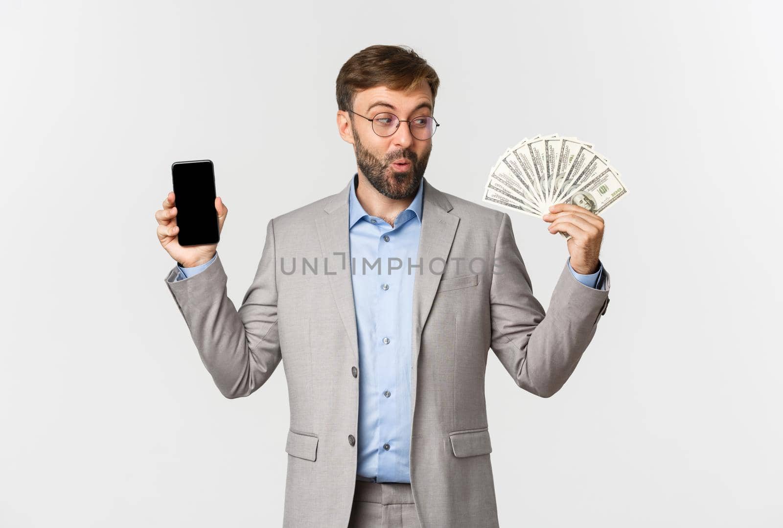 Handsome male boss in grey suit and glasses, smiling pleased as looking at money, showing mobile phone screen, standing over white background.